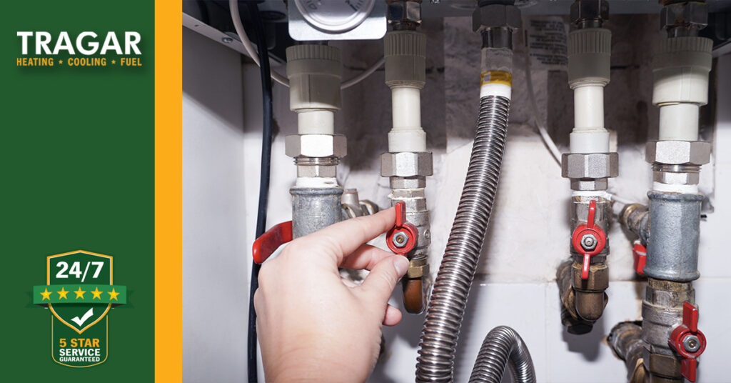 A hand adjusts a red valve on a series of metal pipes under a sink. Tragar Heating logo with a 24/7 service badge is visible on the left.