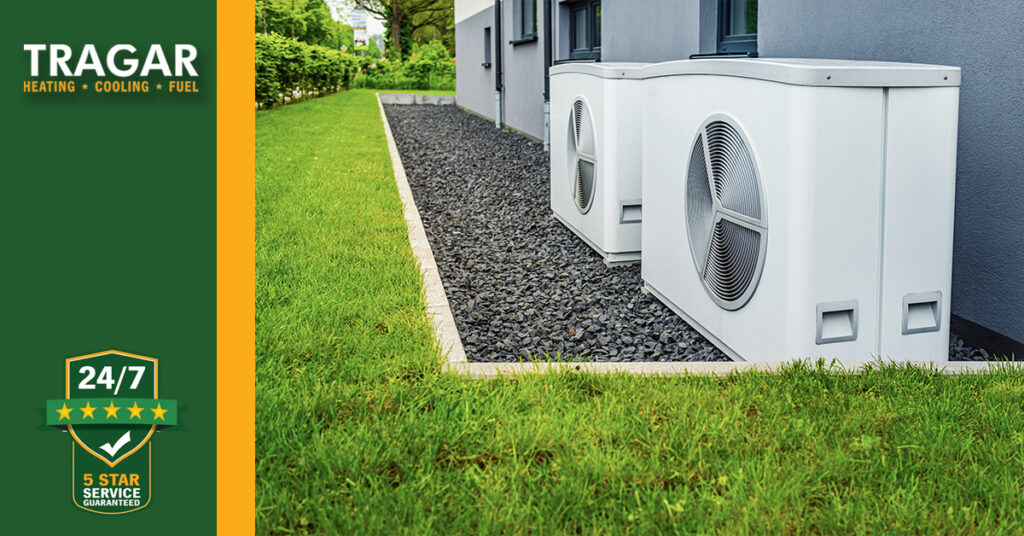 Two outdoor air conditioning units sit on a bed of gravel next to a house. Green grass surrounds the area. The text reads: "TRAGAR Heating • Cooling • Fuel. 24/7 5 Star Service Guaranteed.