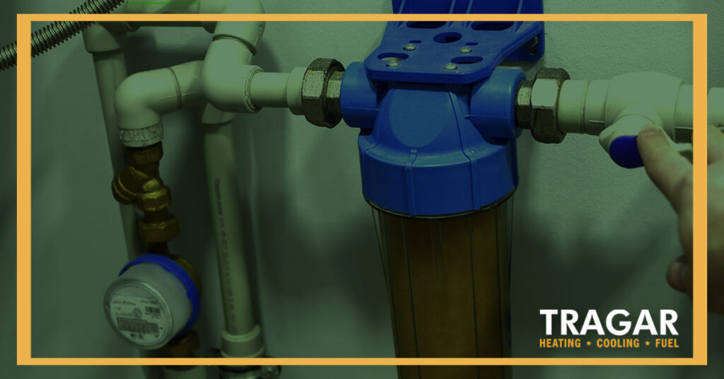 Close-up of a person adjusting a home water filtration system with pipes and a gauge. Tragar logo is visible.