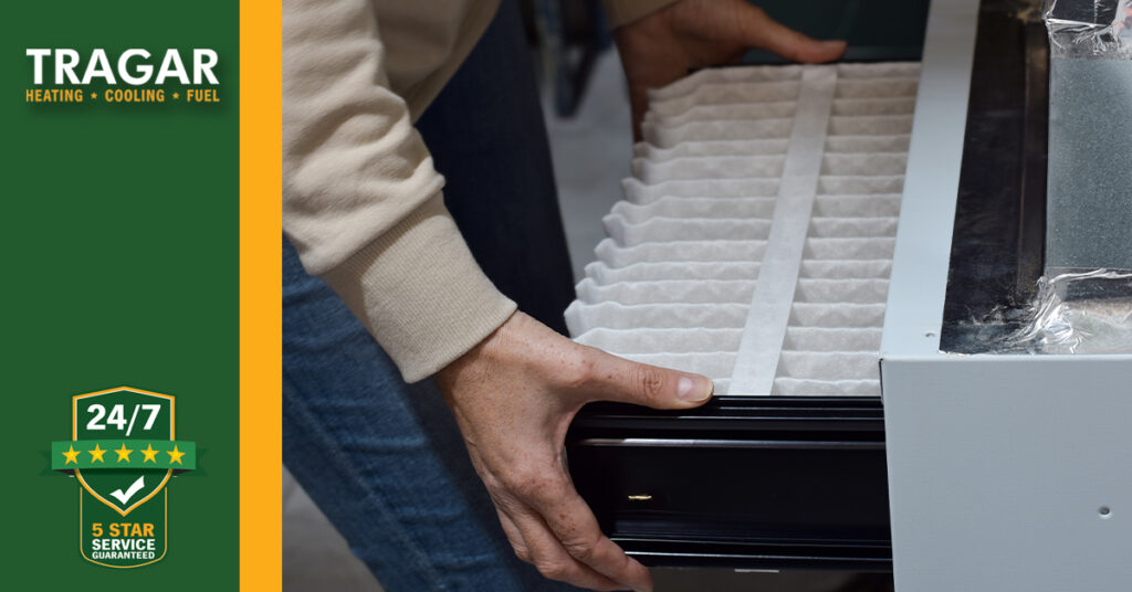 Person replacing a filter in a heating or cooling unit. Tragar logo and 24/7 service badge visible on left side of the image.