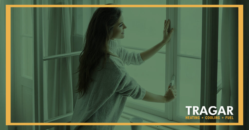 Woman opening a window in a room with sheer curtains, wearing a sweater. Tragar Heating, Cooling, Fuel logo in the corner.