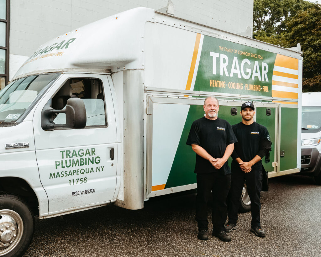 Two men in black uniforms stand beside a Tragar Plumbing service truck parked on a wet street in front of a building, representing the dedicated team you could join—Tragar Plumbing is hiring for rewarding careers.