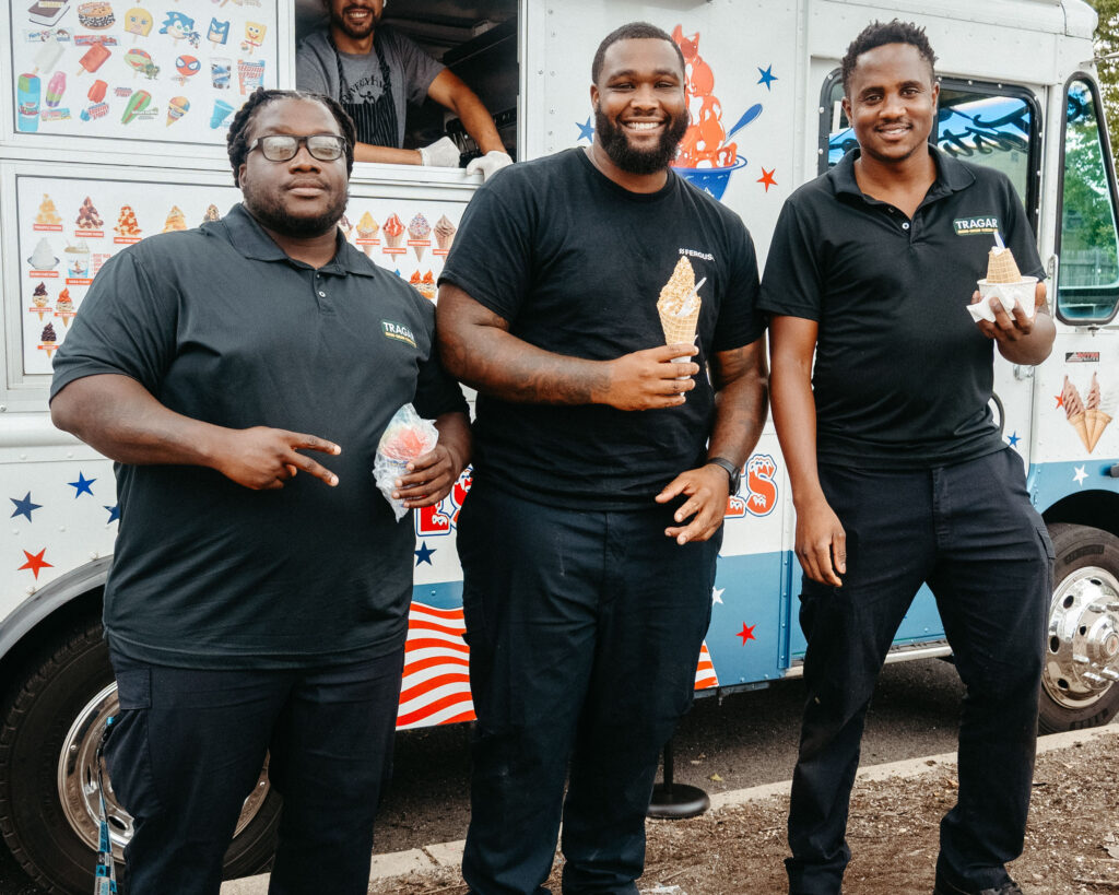 Three men in black polos stand in front of an ice cream truck, each holding an ice cream cone and smiling at the camera, celebrating their rewarding careers with the company.