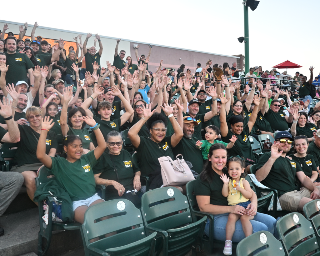 Large group of people in green shirts sitting in stadium seats, smiling and waving at the camera—celebrating new careers and exciting job opportunities.