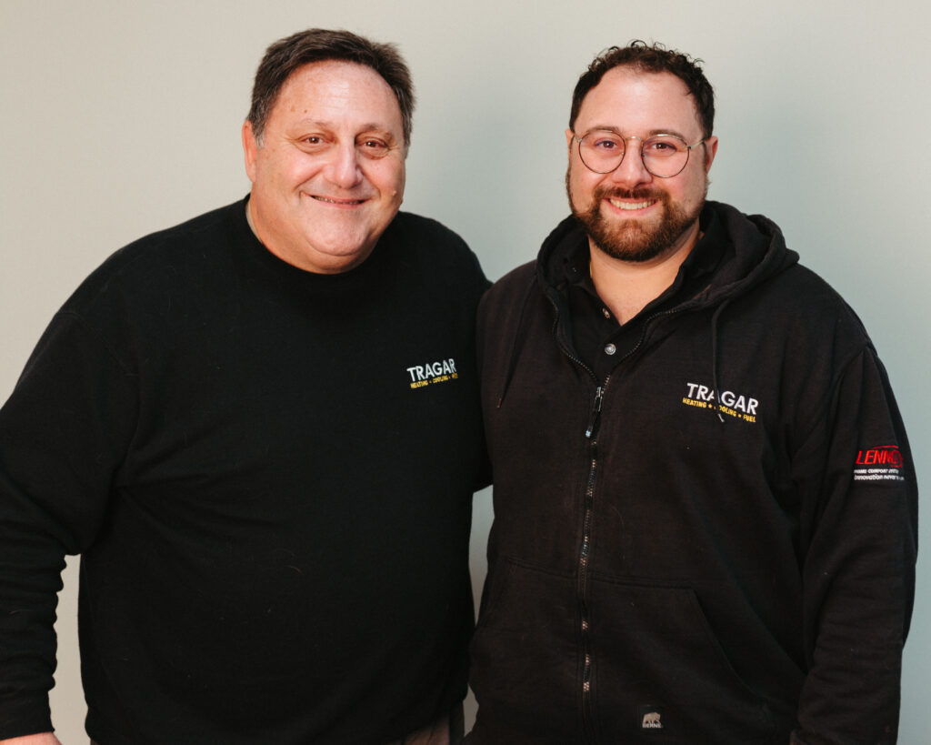 Two men stand side by side, smiling at the camera. Both wear black Tragar-branded shirts, subtly reflecting their pride in the team and its exciting careers. One also wears glasses and a black hoodie. Plain light-colored background.
