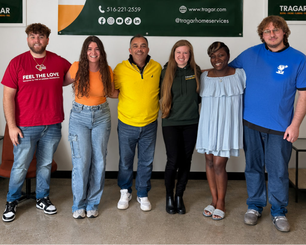 Six people stand indoors, smiling at the camera, in front of green Tragar Home Services signs. Dressed in casual and business casual clothing, they showcase the welcoming team environment that Tragar offers to those interested in careers with a company that is always hiring talented individuals.