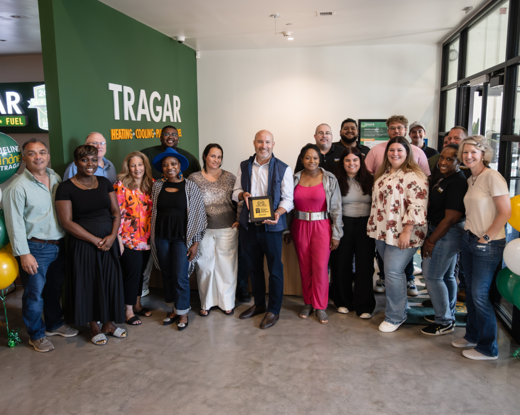 A group of people poses indoors near a green wall with a Tragar sign; one person in the center holds a plaque, reflecting their shared passion for careers and growth.